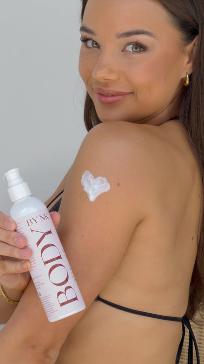 Woman applying sunscreen with a bottle labeled 'BODY' on a plain background